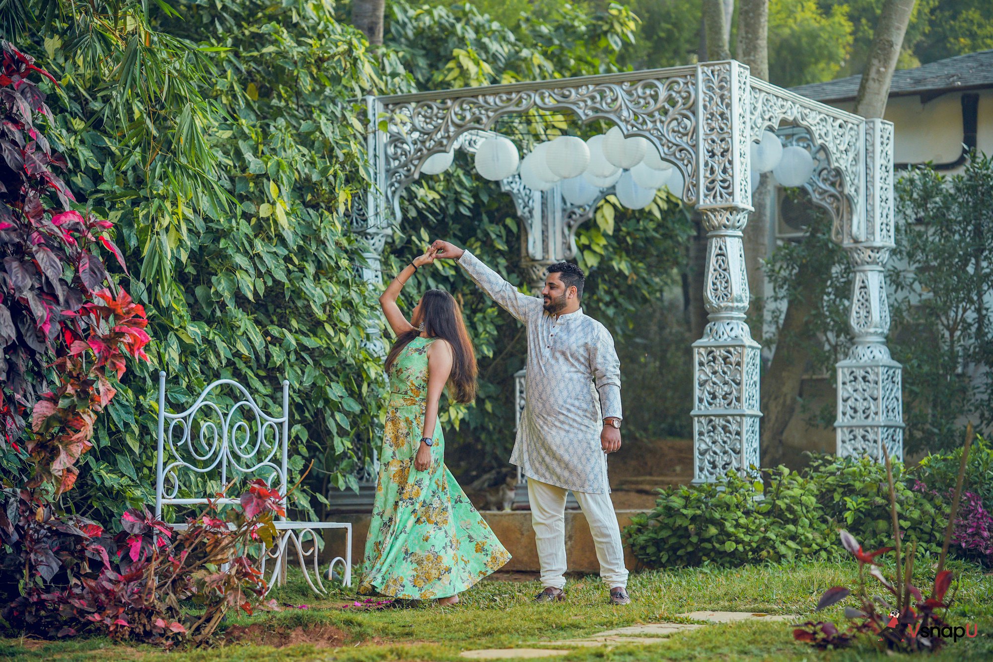 Pre-wedding photo of an ethnic couple dancing in a garden, with a charming bench set against a backdrop of greenery.